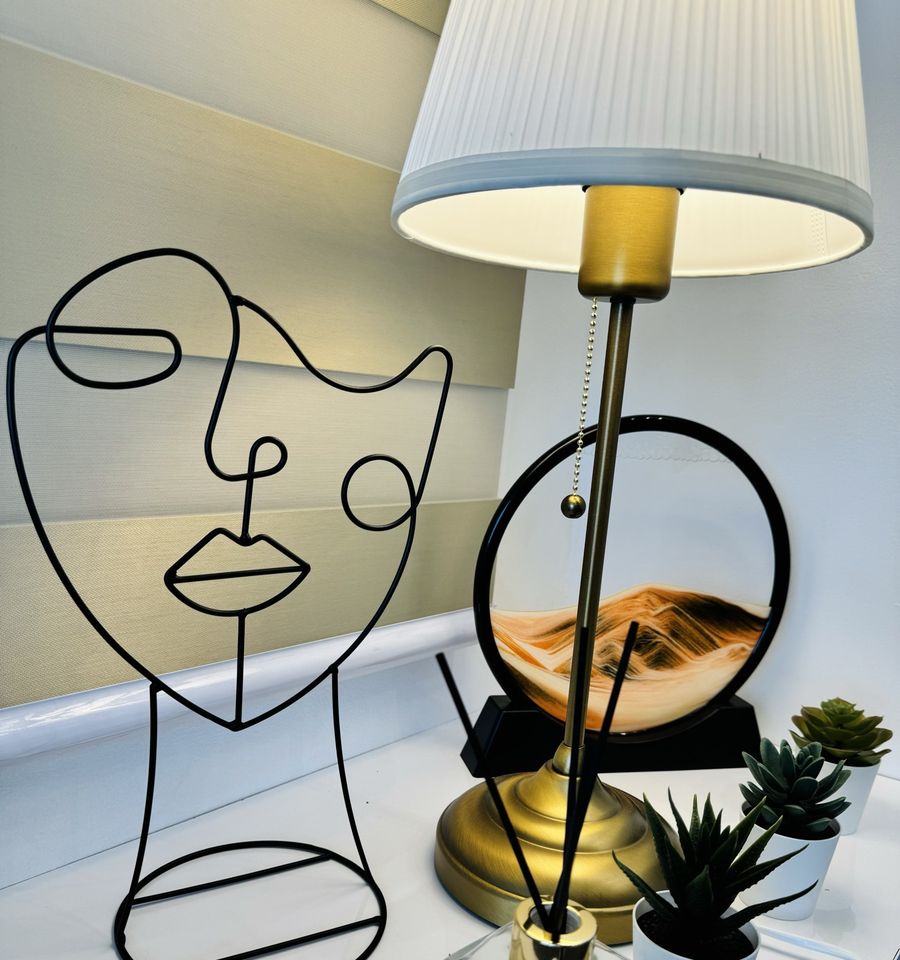Line-art face sculpture, a gold table lamp and small succulents on a white counter